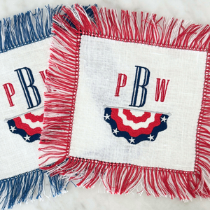 White Linen Cocktail Napkins with Blue and Red Fringe shown with a Red, white and Blue Stars and Stripes Bunting and and Red and Blue Monogram.