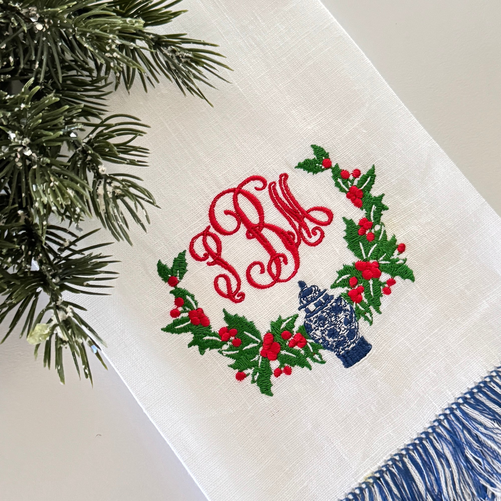 Holly swag embroidery design with a chinoiserie blue and white ginger jar. Shown on a blue and white linen fringe guest towel.