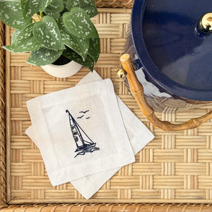 A stack of square white linen cocktail napkins featuring a navy blue embroidered sailboat design displayed on a woven rattan tray with a green plant and blue ice bucket with bamboo handle in the background.