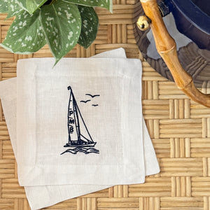 A stack of square white linen cocktail napkins featuring a navy blue embroidered sailboat design displayed on a woven rattan tray with a green plant and blue ice bucket with bamboo handle in the background.
