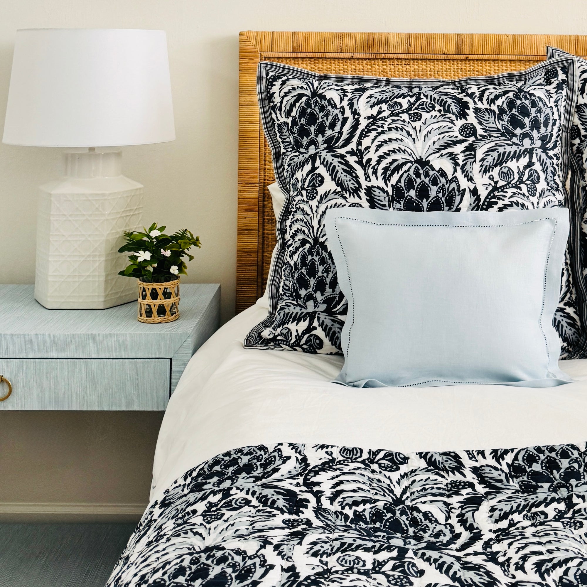 Sky Blue Linen boudoir pillow featured on a bed with a Serena and Lily rattan headboard and blue and white Serena and Lily artichoke print bedding. There is also a light blue nightstand, a white table lamp and a small bouquet of flowers in a wicker covered vase.